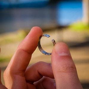 May include: A silver ring with a textured surface, held open between two fingers.