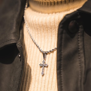 May include: A silver cross pendant necklace with an intricate design hangs against a beige turtleneck jumper. The necklace is on a rope chain. A black leather jacket is visible.