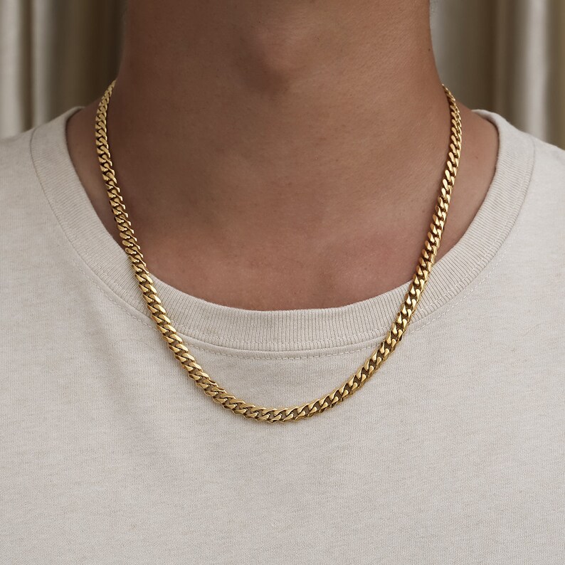 May include: A gold chain necklace with a curb link style. The chain is worn by a person with a light beige shirt.