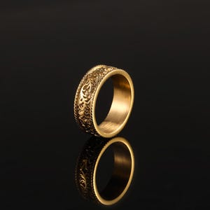May include: A gold-toned band ring featuring an intricate, embossed floral or vine-like pattern covering the outer surface. The ring has a polished finish and a smooth, plain interior, reflecting clearly against the dark, reflective surface below.