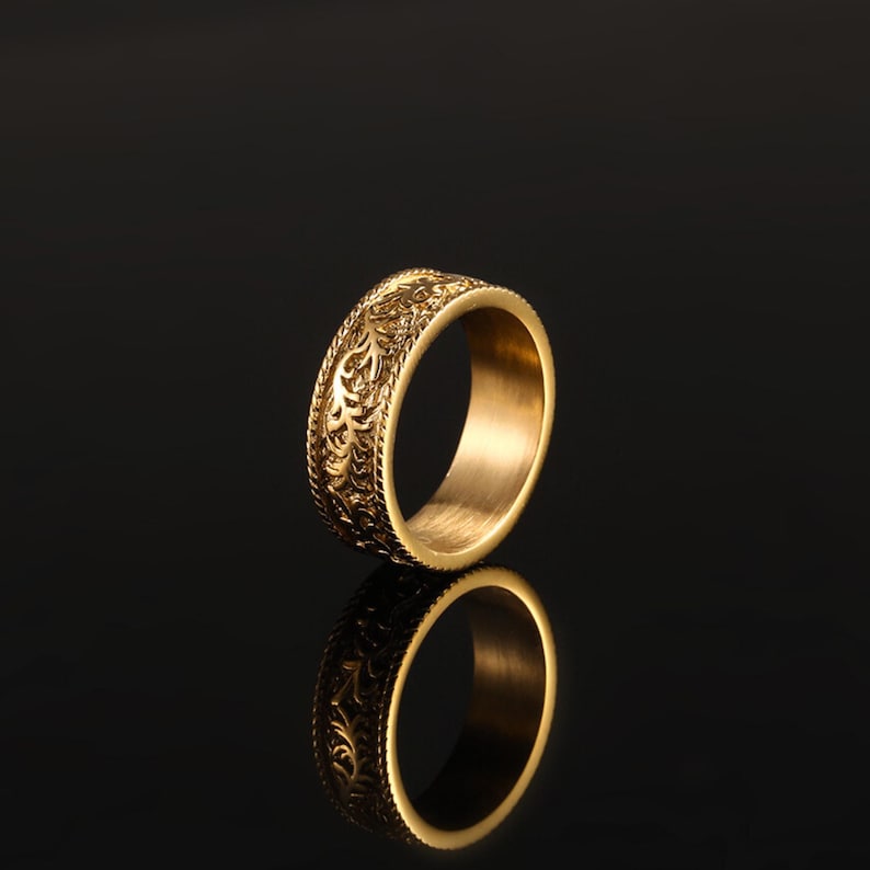 May include: A gold-toned band ring featuring an intricate, embossed floral or vine-like pattern covering the outer surface. The ring has a polished finish and a smooth, plain interior, reflecting clearly against the dark, reflective surface below.