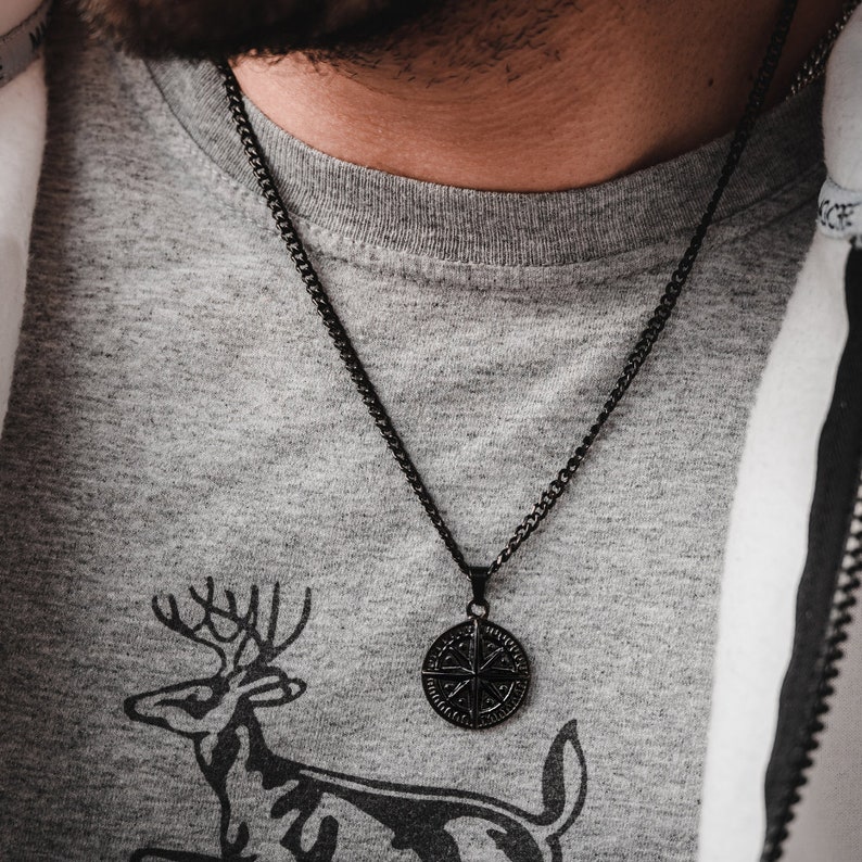 K&ouml;nnte beinhalten: Eine schwarze Kettenkette mit einem Kompass-Anh&auml;nger. Der Anh&auml;nger ist rund und hat ein detailliertes Design. Die Kette wird &uuml;ber einem grauen T-Shirt mit einem Hirsch-Motiv getragen.
