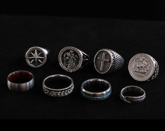 May include: A collection of eight silver rings with various designs, including a compass, a cross, a saint, and a dragon. The rings are arranged on a black surface.