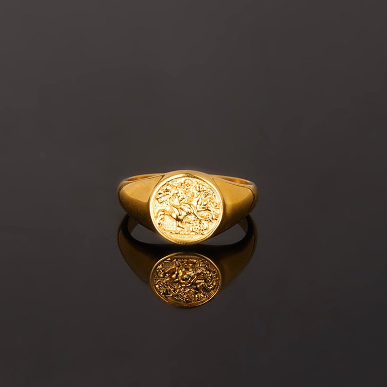 May include: A gold signet ring featuring a circular face with an embossed design of a figure on horseback. The ring has a polished, smooth band that tapers toward the back, reflecting clearly on the dark surface beneath the jewelry piece.
