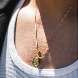 May include: Gold-colored pendant necklace featuring an oval pendant with a religious figure and a small cross. The necklace is worn over a white tank top. The chain and pendant appear to be made of a shiny metal.