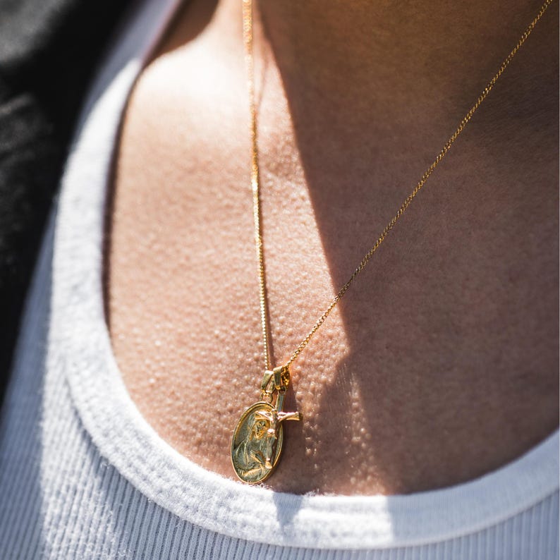 May include: Gold-colored pendant necklace featuring an oval pendant with a religious figure and a small cross. The necklace is worn over a white tank top. The chain and pendant appear to be made of a shiny metal.
