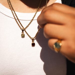 May include: Gold-toned necklaces with square pendants, one green and one red, hang against a white shirt. A gold ring with a green stone is visible on a hand. The jewelry is the focal point, showcasing a minimalist aesthetic.