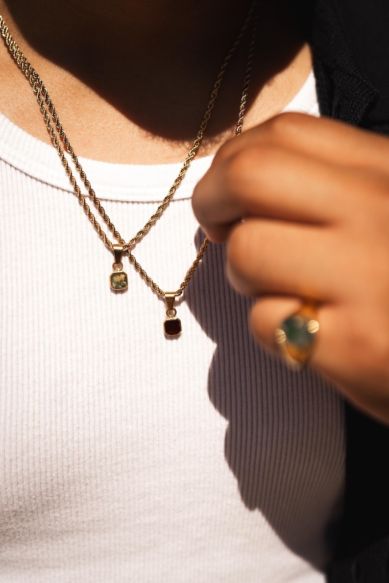 May include: Gold-toned necklaces with square pendants, one green and one red, hang against a white shirt. A gold ring with a green stone is visible on a hand. The jewelry is the focal point, showcasing a minimalist aesthetic.