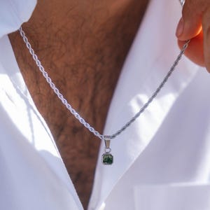 Op de afbeelding: Zilverkleurige touwketting met een kleine, vierkante groene hanger. De hanger heeft een donkergroene, getextureerde uitstraling. De ketting wordt getoond tegen een wit overhemd.