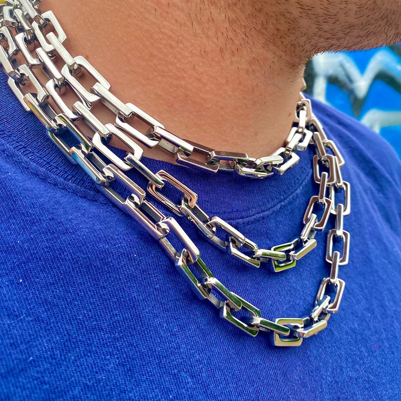 May include: Three silver-colored chain necklaces with rectangular links are displayed. The chains are layered, creating a bold, eye-catching accessory. The background is a blue shirt and a blurred wall.