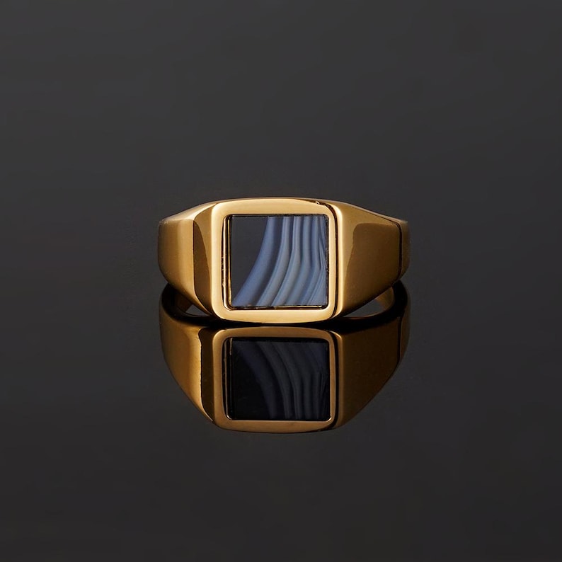 May include: A gold-toned signet ring featuring a square-shaped setting. The center of the ring contains a flat, smooth inlay of blue-grey banded agate stone with distinct, parallel white and dark grey stripes running vertically across the surface.