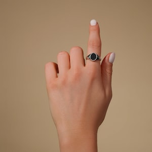 Op de afbeelding: Een zilveren zegelring met een zwarte ovale steen. De ring zit aan een vinger, met de hand omhoog tegen een neutrale achtergrond. Het ontwerp van de ring is eenvoudig en klassiek.