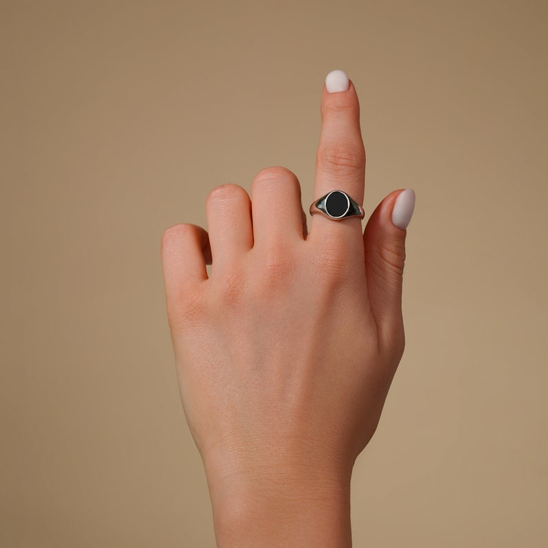 Op de afbeelding: Een zilveren zegelring met een zwarte ovale steen. De ring zit aan een vinger, met de hand omhoog tegen een neutrale achtergrond. Het ontwerp van de ring is eenvoudig en klassiek.