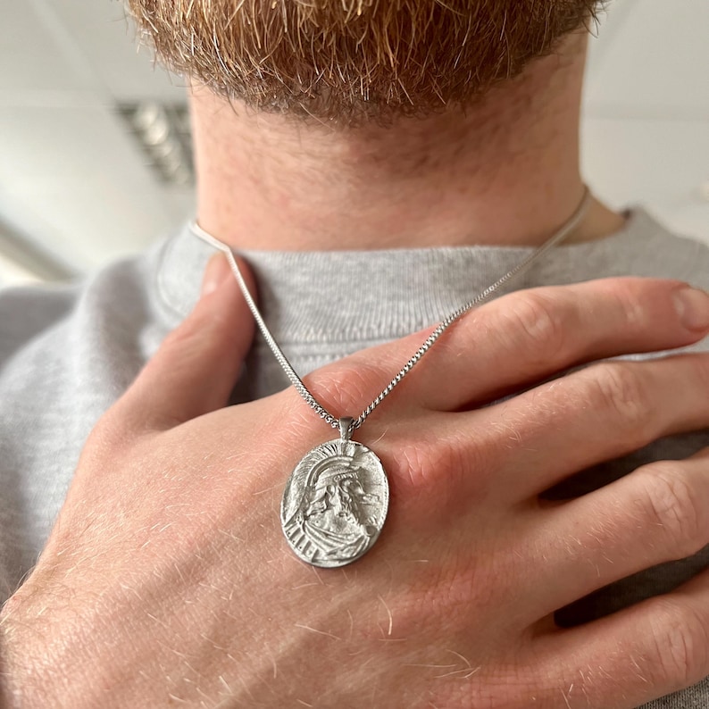 May include: A silver chain necklace with a silver oval pendant featuring a detailed profile of a person's face.