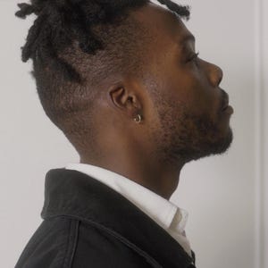 Puede incluir: Retrato de perfil de una persona con piel oscura, pelo corto y un peque&ntilde;o pendiente de aro dorado. Llevan una chaqueta negra sobre una camisa blanca con cuello. El fondo es una pared blanca lisa.