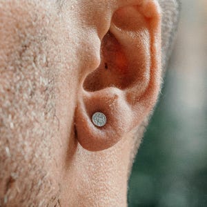 Può includere: Un piccolo orecchino a lobo circolare color argento con una superficie testurizzata e granulosa. L'orecchino è posizionato al centro del lobo, caratterizzato da una lucentezza metallica e un profilo piatto a forma di disco sulla pelle.