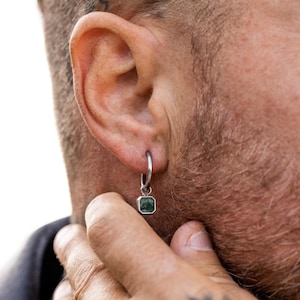 Può includere: Un orecchino a cerchio d'argento con un pendente quadrato di pietra preziosa verde scuro. L'orecchino è indossato sull'orecchio. La pietra preziosa è incastonata in una lunetta d'argento. La persona ha capelli corti e scuri e barba.