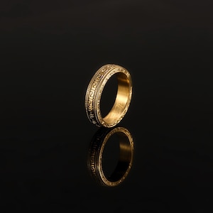 May include: A gold-toned metal ring featuring a continuous Greek key pattern engraved around the exterior band. The ring has a polished finish and a smooth interior surface, reflecting light to highlight the intricate geometric design details.