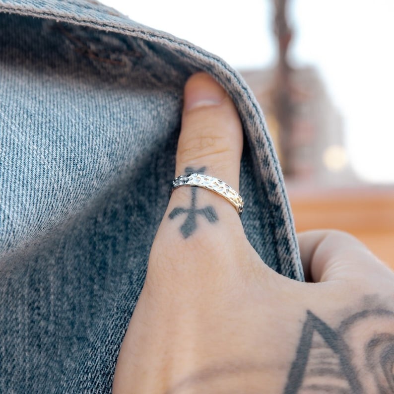 May include: A silver hammered ring worn on a finger with a cross tattoo.