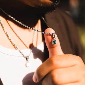 May include: Silver-toned necklace with a square, emerald-green pendant. The pendant is held up by a finger, showcasing the jewelry's design. A second necklace with a white square pendant is also visible.