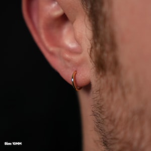 Puede incluir: Un pendiente de aro dorado, de 10 mm, se muestra en la oreja de una persona. El pendiente tiene un dise&ntilde;o circular simple con un acabado pulido. El fondo es oscuro, enfatizando la forma y el color del pendiente.
