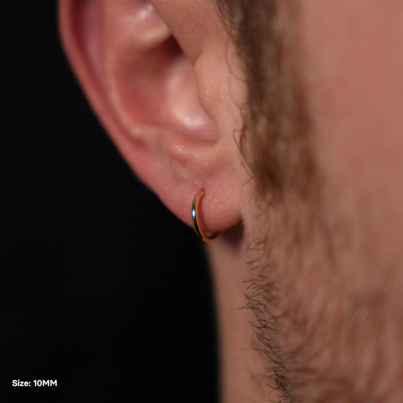 Puede incluir: Un pendiente de aro dorado, de 10 mm, se muestra en la oreja de una persona. El pendiente tiene un dise&ntilde;o circular simple con un acabado pulido. El fondo es oscuro, enfatizando la forma y el color del pendiente.