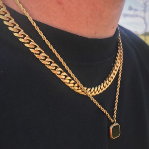 May include: Gold-toned necklace set featuring a thick curb chain, a rope chain, and a pendant necklace with a black square stone. The necklaces are layered against a black shirt.