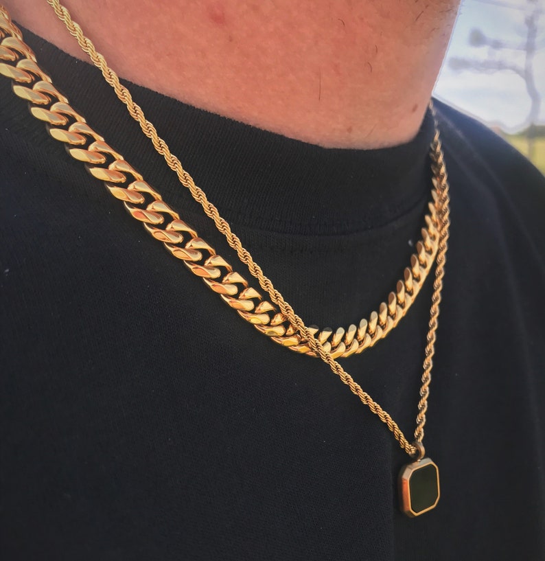 May include: Gold-toned necklace set featuring a thick curb chain, a rope chain, and a pendant necklace with a black square stone. The necklaces are layered against a black shirt.