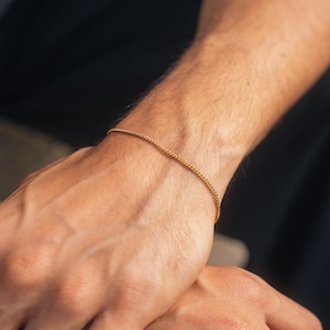 Könnte beinhalten: Ein filigranes, goldfarbenes Armband mit Perlen. Das Armband wird am Handgelenk getragen, wobei Arm und Hand im Vordergrund stehen. Der Hintergrund ist dunkel und hebt den Schmuck hervor. Das Armband besteht aus kleinen, runden Perlen, die aufgefädelt sind.