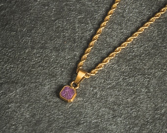 May include: A gold-colored necklace with a square, purple pendant. The pendant is set in a gold frame and hangs from a twisted rope chain. The necklace is displayed on a dark gray surface.