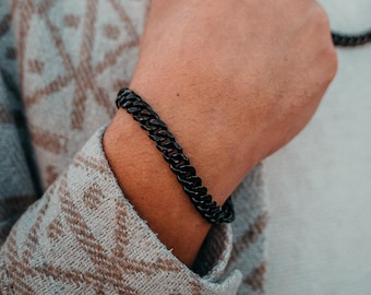Puede incluir: Un brazalete de cadena negra con un diseño de eslabones de curb. El brazalete se lleva en la muñeca.