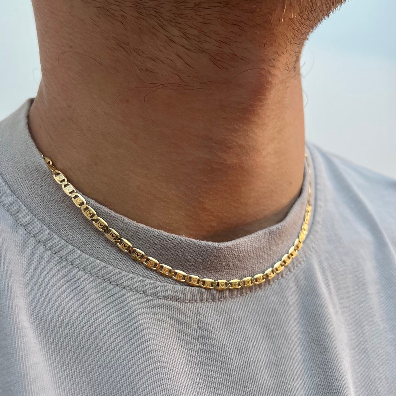 Może przedstawiać: Złoty naszyjnik z łańcuszkiem w stylu marynarskim. Łańcuszek nosi osoba w jasnoszarym t-shirt.