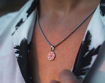 May include: A silver necklace with a geometric pendant featuring a red and white design. The pendant is hexagonal and hangs from a silver chain. The person is wearing a white shirt with a black and white pattern.