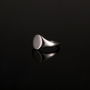 May include: A silver signet ring with a smooth, oval-shaped top. The ring is on a black background.