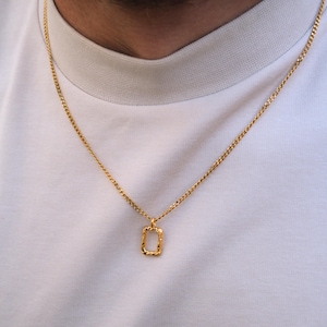 May include: A gold-colored necklace with a rectangular pendant. The pendant has a textured surface and is suspended from a delicate chain. The necklace is worn against a white t-shirt.