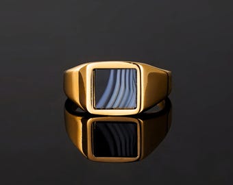 May include: A gold-colored signet ring with a square face. The face of the ring features a rectangular inlay with a blue and white striped pattern. The ring is displayed on a reflective black surface.