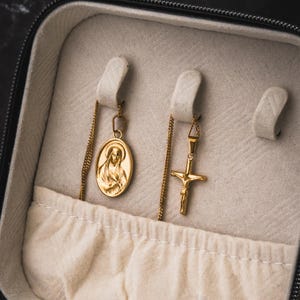 May include: Gold-toned necklaces displayed in a jewelry case. One necklace features an oval pendant with a religious figure, and the other has a cross pendant. The case is lined with a light-colored fabric.