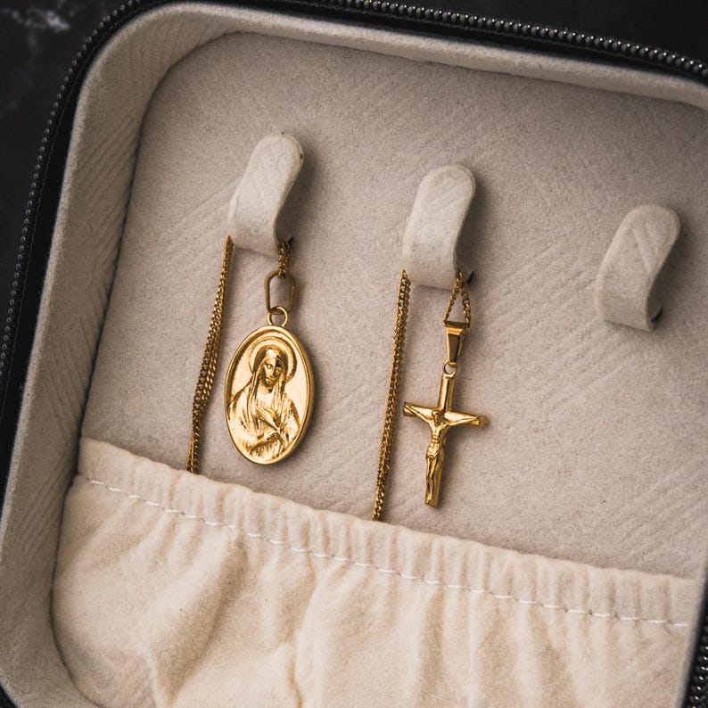 May include: Gold-toned necklaces displayed in a jewelry case. One necklace features an oval pendant with a religious figure, and the other has a cross pendant. The case is lined with a light-colored fabric.
