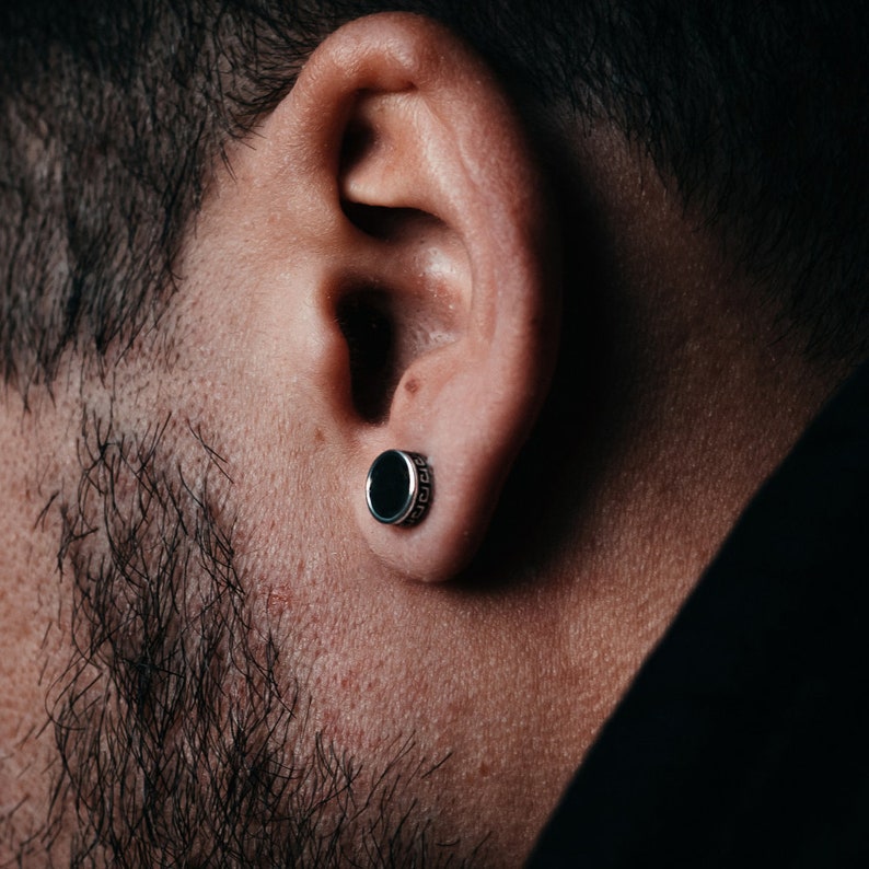 May include: A close-up of a person's ear with a black and silver stud earring. The earring has a round, black stone and a silver band with a decorative pattern.