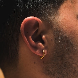 Puede incluir: Primer plano de un pendiente de aro dorado con superficie texturizada. El pendiente se lleva en la oreja de una persona con cabello oscuro y barba. El pendiente tiene un diseño circular pequeño.