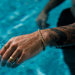 May include: A gold rope bracelet and two gold rings adorn a hand submerged in blue water. One ring has a black stone, the other is a simple signet style. The arm has a black tattoo.