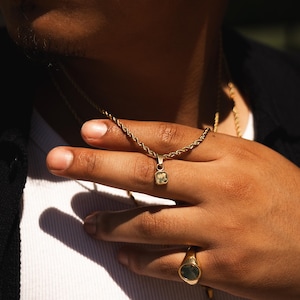 May include: Gold-toned necklace with a small, square pendant featuring a light green gemstone. The necklace is displayed on a hand, along with a matching gold ring with a dark green stone. The jewelry is set against a dark background.