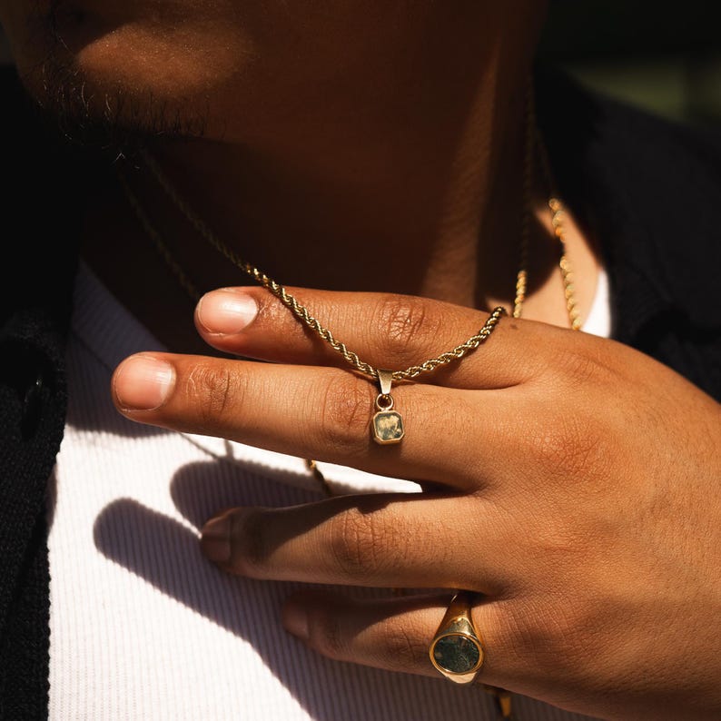 May include: Gold-toned necklace with a small, square pendant featuring a light green gemstone. The necklace is displayed on a hand, along with a matching gold ring with a dark green stone. The jewelry is set against a dark background.