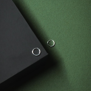 May include: Two small, silver hoop earrings. One earring is placed on the corner of a black box, and the other is on a green fabric background. The earrings are simple and minimalist in design.