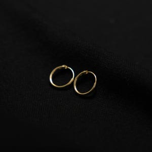 Puede incluir: Un par de pendientes de aro peque&ntilde;os, de color dorado. Los pendientes son circulares y parecen estar hechos de un metal fino. Se colocan sobre un fondo oscuro y texturizado, creando un contraste que resalta su forma y color.