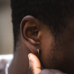 Op de afbeelding: Close-up van een oor met een kleine gouden oorring. Het oor is bruin en de persoon heeft kort zwart haar.