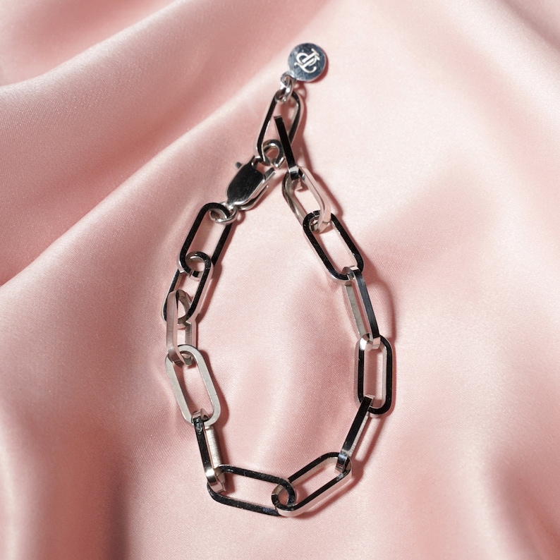 May include: A silver chain link bracelet with a lobster clasp and a small circular charm. The bracelet is displayed on a pink satin fabric, creating a contrast between the shiny metal and the soft background.
