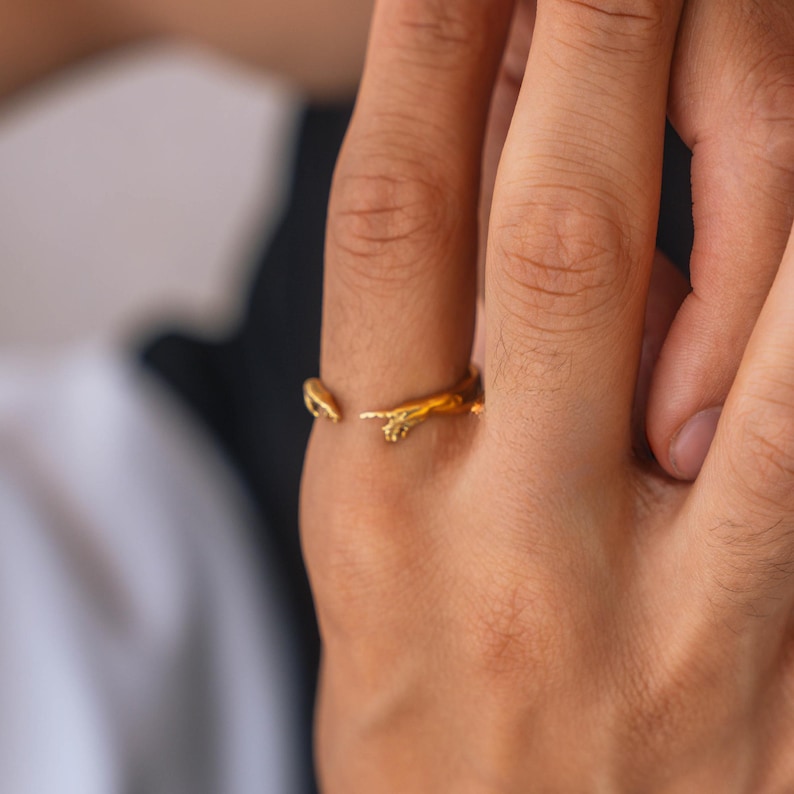 May include: A gold-toned open ring featuring two sculpted hands reaching toward each other, designed to mimic the iconic artistic depiction of fingers nearly touching. The band is crafted with a polished metallic finish and an adjustable open gap.