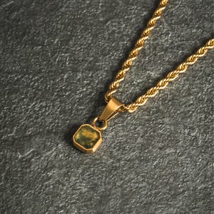 May include: Gold-toned necklace with a square pendant featuring a green and gold stone. The chain has a rope design. The pendant is suspended from a small bail. The necklace is displayed on a gray surface.