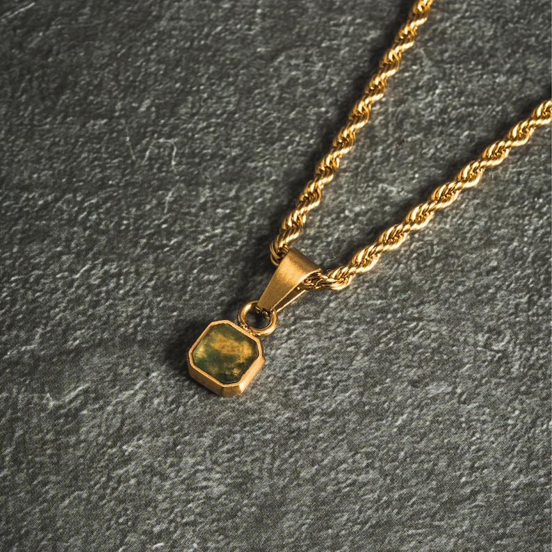 May include: Gold-toned necklace with a square pendant featuring a green and gold stone. The chain has a rope design. The pendant is suspended from a small bail. The necklace is displayed on a gray surface.